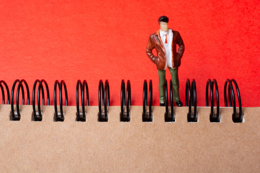 Small male figurine standing with hands in pockets on a spiral-bound notebook against a bright red background.