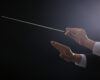 Conductor's hands guiding with a baton mid-stroke against a dark background.