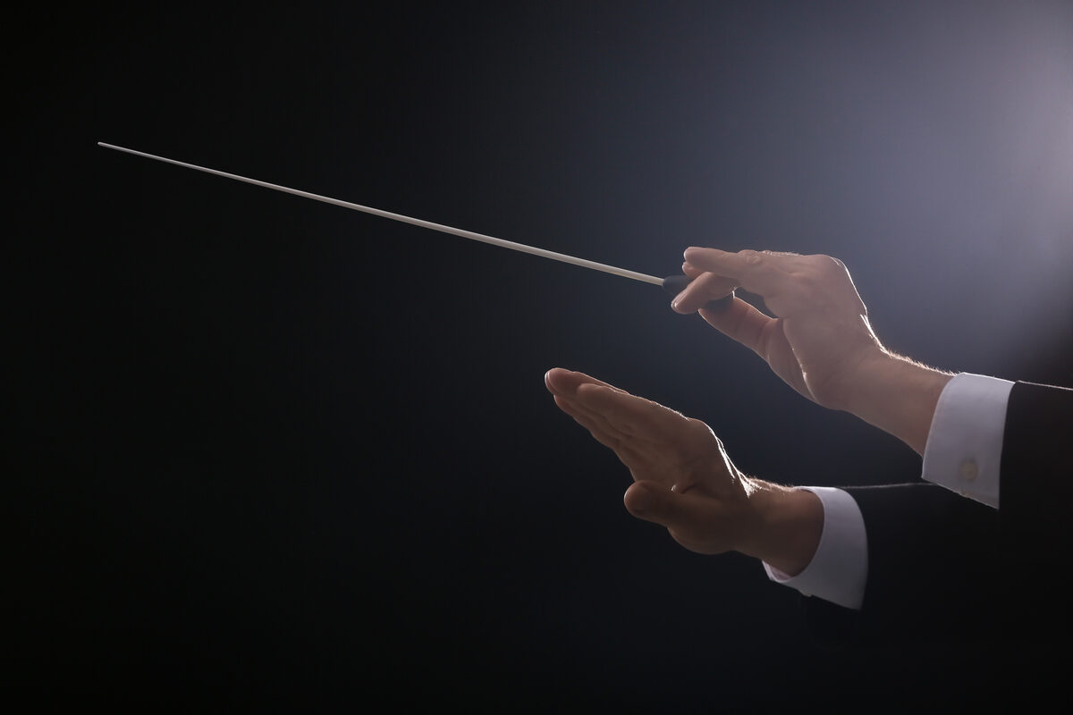Conductor's hands guiding with a baton mid-stroke against a dark background.