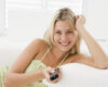 Smiling blonde woman lying on a white sofa, holding a TV remote at home.