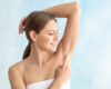 Smiling woman lifts her left arm to reveal her bare underarm against a light blue background, suggesting armpit care or grooming.