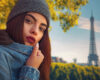 Young woman in a gray beanie and denim jacket posing outdoors with the Eiffel Tower in the background.
