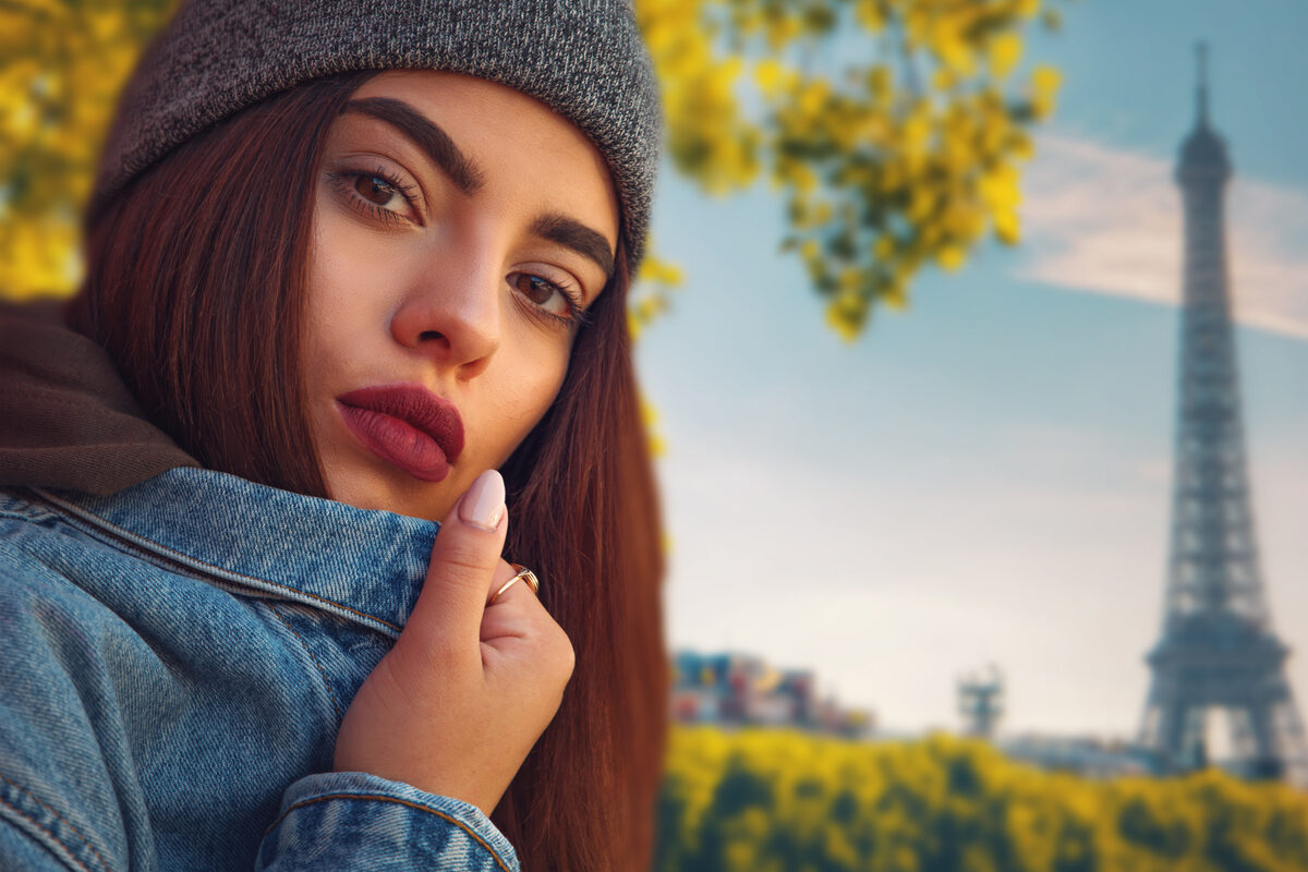 Young woman in a gray beanie and denim jacket posing outdoors with the Eiffel Tower in the background.