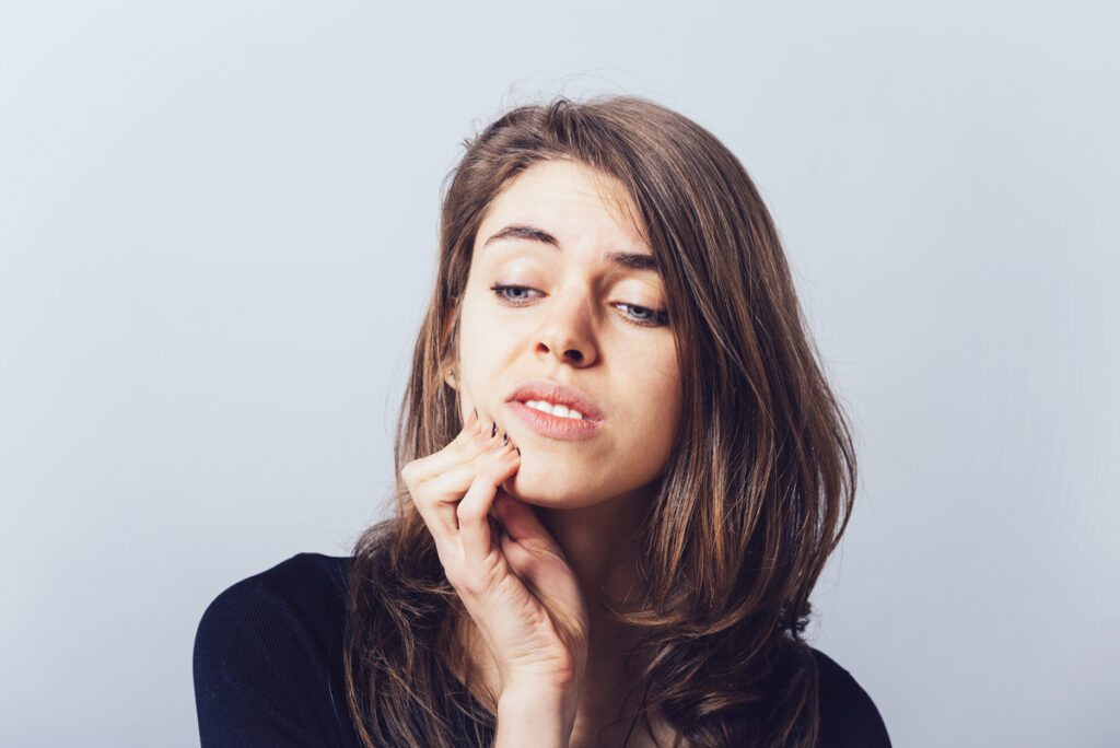 Woman with long brown hair in a dark top touches her jaw, looking to the side in a thoughtful pose.