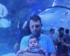 Man in a light blue shirt holding a baby inside a transparent aquarium tunnel, with blue lighting and fish swimming around.