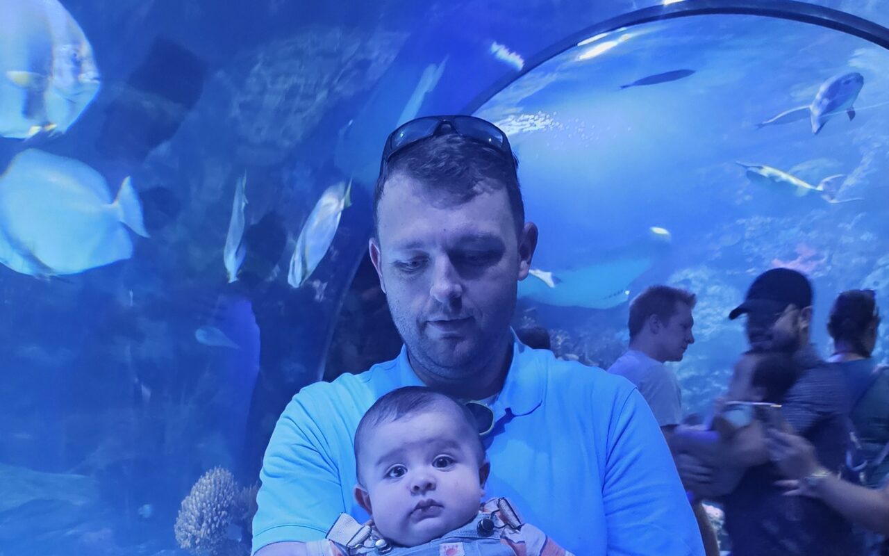 Man in a light blue shirt holding a baby inside a transparent aquarium tunnel, with blue lighting and fish swimming around.