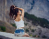 Young woman in a white tank top and denim shorts sits on a concrete ledge, tying her hair, with rugged cliffs in the background.