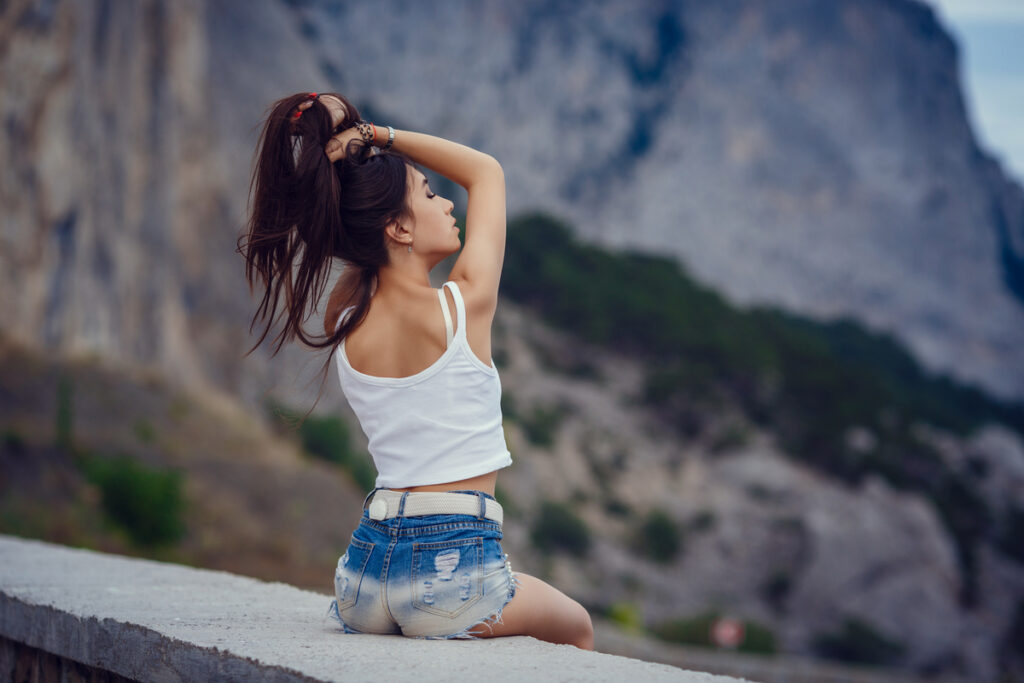 Young woman in a white tank top and denim shorts sits on a concrete ledge, tying her hair, with rugged cliffs in the background.