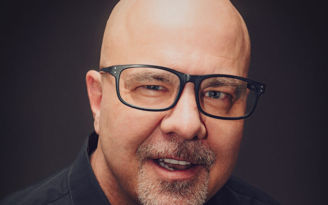 Portrait of a bald man wearing black-framed glasses, smiling softly against a dark background.