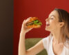 Woman about to bite into a hamburger, smiling, indoors against a red wall with a lettuce-topped burger in hand.