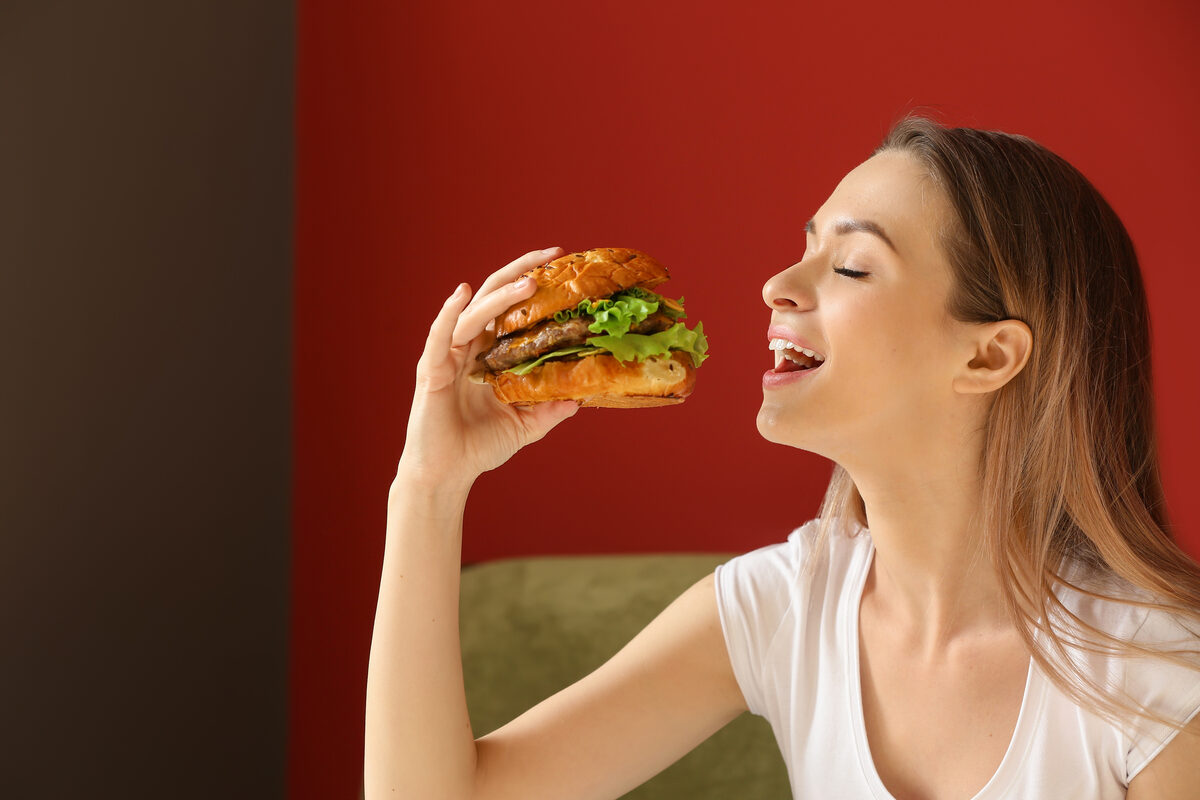 Woman about to bite into a hamburger, smiling, indoors against a red wall with a lettuce-topped burger in hand.