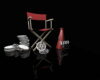 Film production gear: red director's chair, stacked movie reels, a clapperboard, and a red studio light on a reflective black surface.