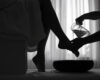Two silhouetted figures: one person holds a foot while another pours water from a pitcher into a basin, performing a foot-washing scene in a dim room.
