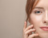 Close-up of a woman's face as she lightly rests fingers on her cheek, with soft makeup and a neutral background.