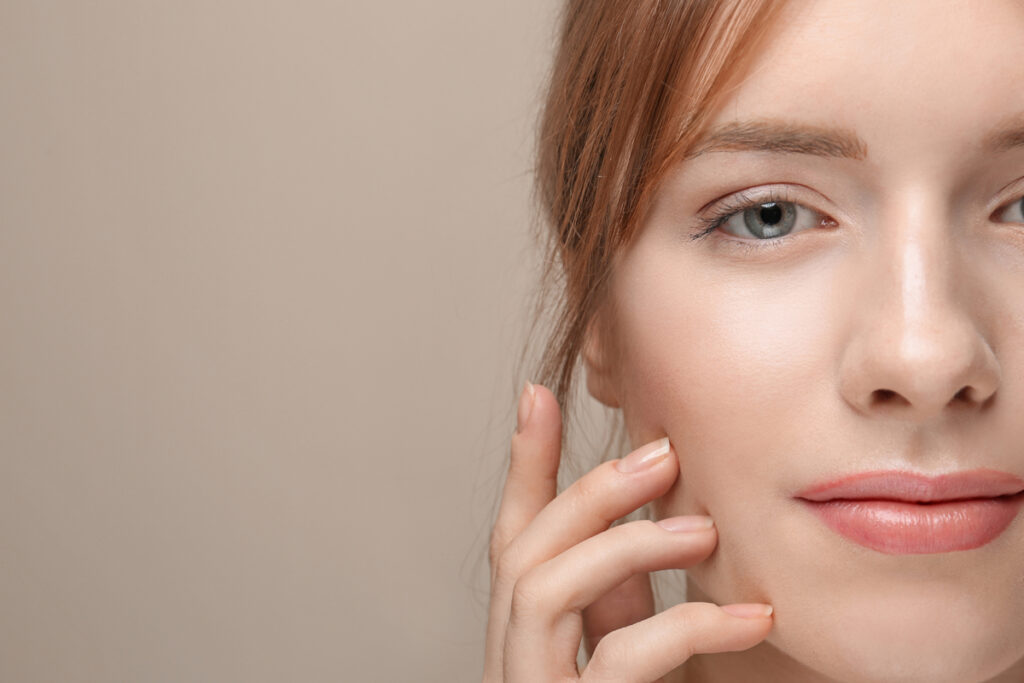 Close-up of a woman's face as she lightly rests fingers on her cheek, with soft makeup and a neutral background.