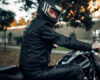 Motorcyclist in a black helmet and leather jacket seated on a motorcycle in a park, looking over their shoulder.