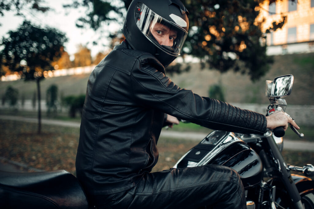 Motorcyclist in a black helmet and leather jacket seated on a motorcycle in a park, looking over their shoulder.