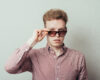 Young man in a red checkered shirt pulls down sunglasses to look at the camera, with a neutral expression.