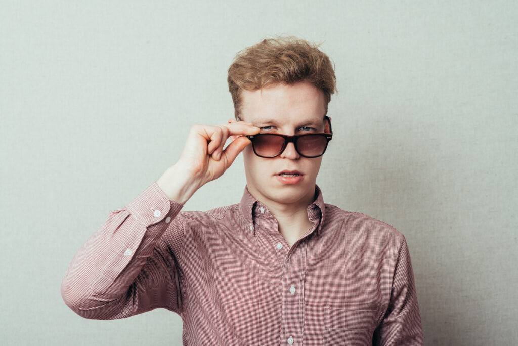 Young man in a red checkered shirt pulls down sunglasses to look at the camera, with a neutral expression.
