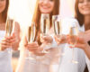 People clinking champagne flutes in a celebratory toast, smiling in white outfits under warm light.