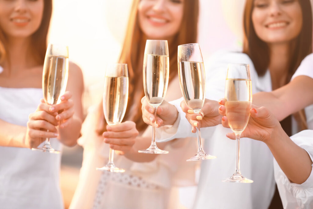 People clinking champagne flutes in a celebratory toast, smiling in white outfits under warm light.