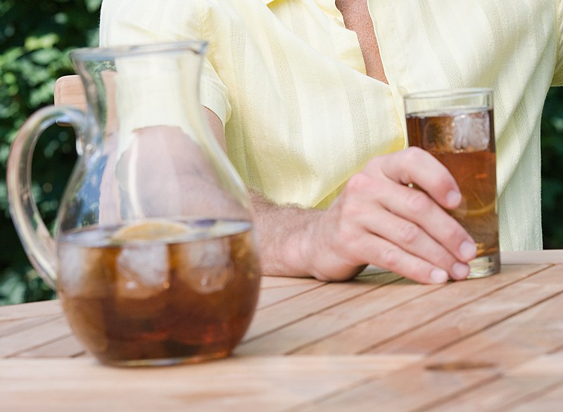 Person in a pale yellow shirt sits at a wooden outdoor table with a pitcher and a glass of iced tea nearby on the table.
