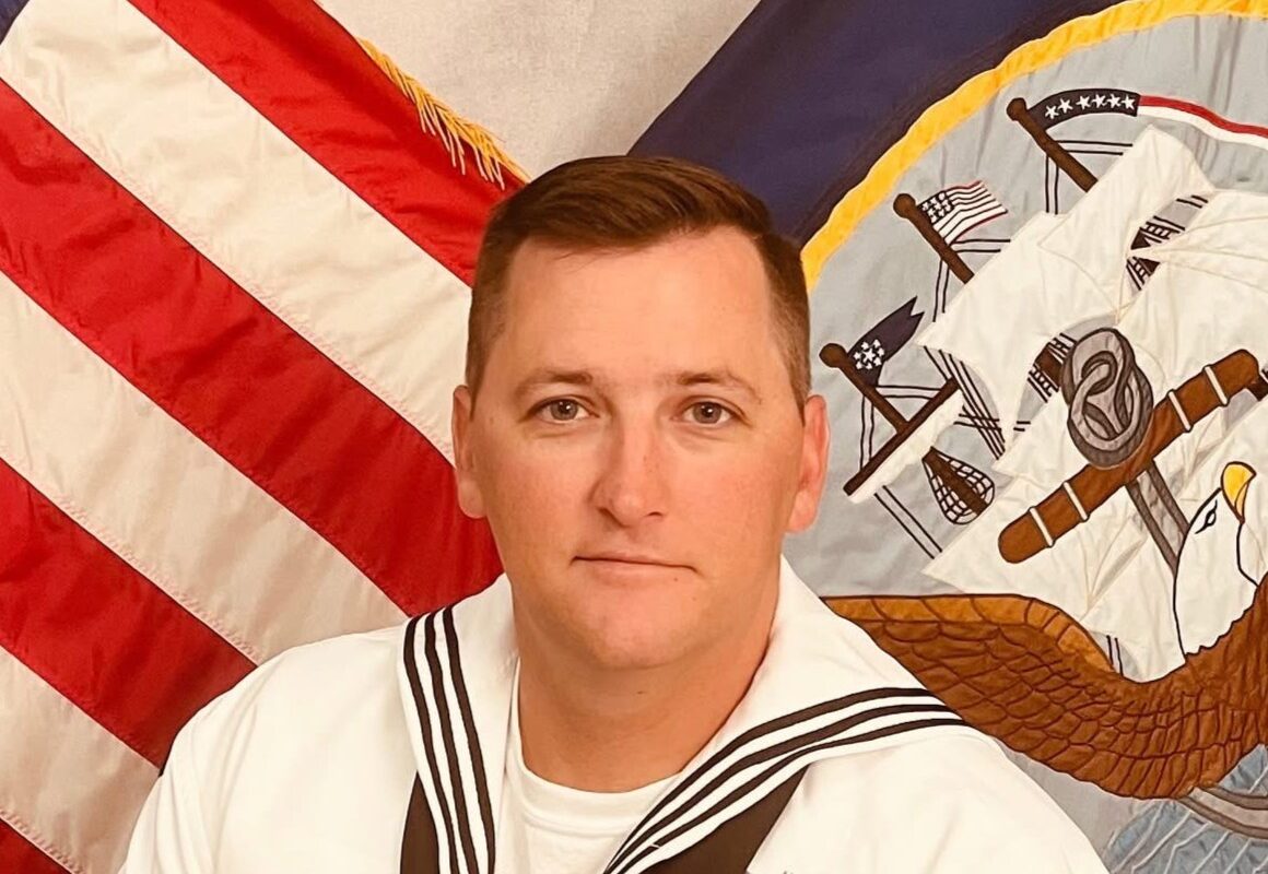 Sailor in white dress uniform with ribbons sits before a US flag and a flag with an eagle emblem in a ceremonial setting.