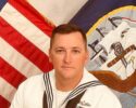 Sailor in white dress uniform with ribbons sits before a US flag and a flag with an eagle emblem in a ceremonial setting.