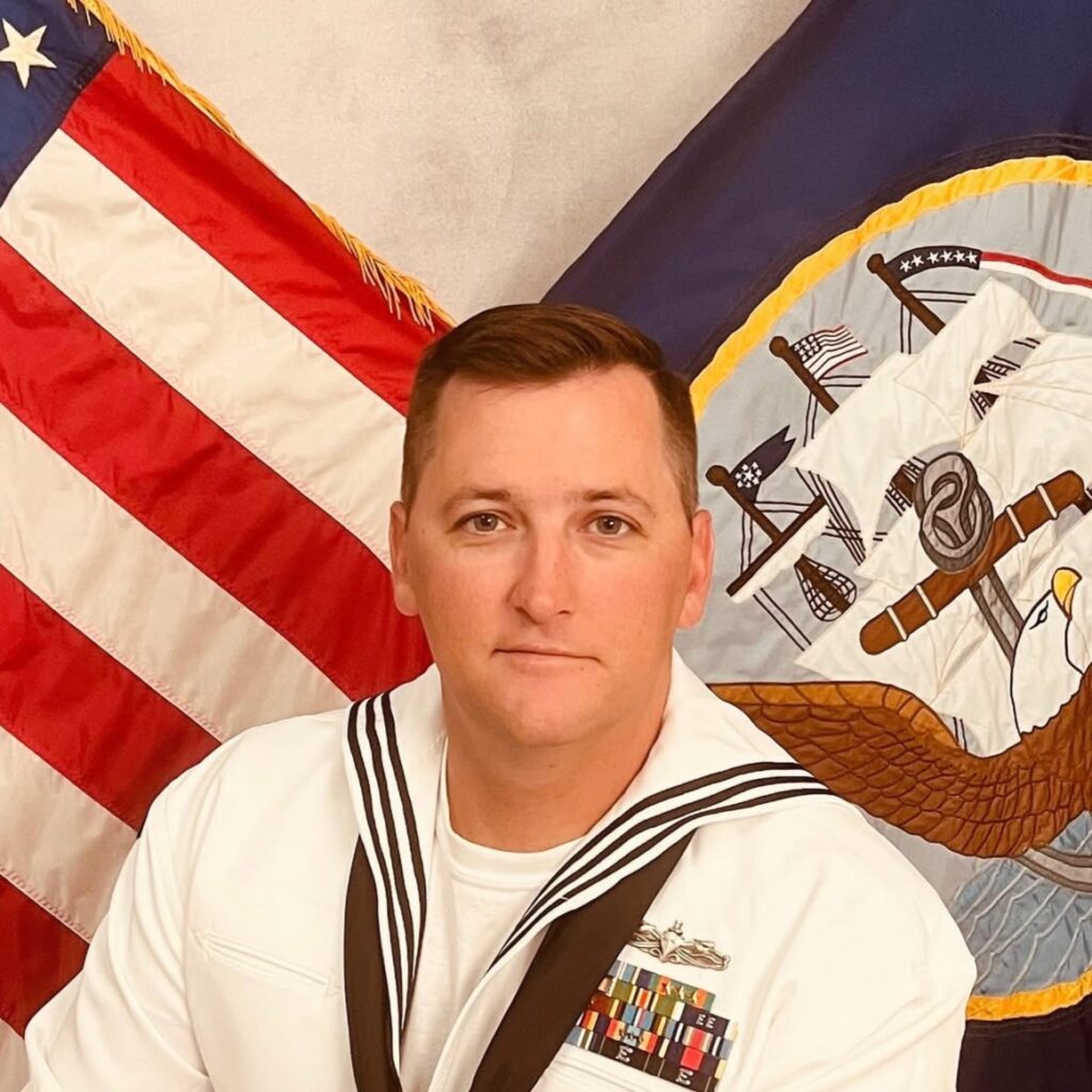 Sailor in white dress uniform with ribbons sits before a US flag and a flag with an eagle emblem in a ceremonial setting.