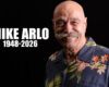 Memorial portrait of Mike Arlo, 1948–2026, smiling in a blue tropical shirt against a black background.