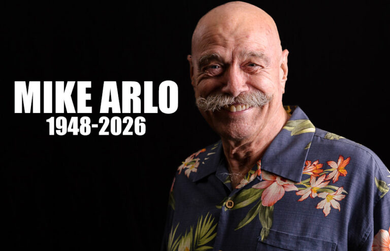 Memorial portrait of Mike Arlo, 1948–2026, smiling in a blue tropical shirt against a black background.