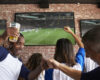 Group of friends in a sports bar celebrating a soccer match as they fist-bump and cheer at big screens on a brick wall.