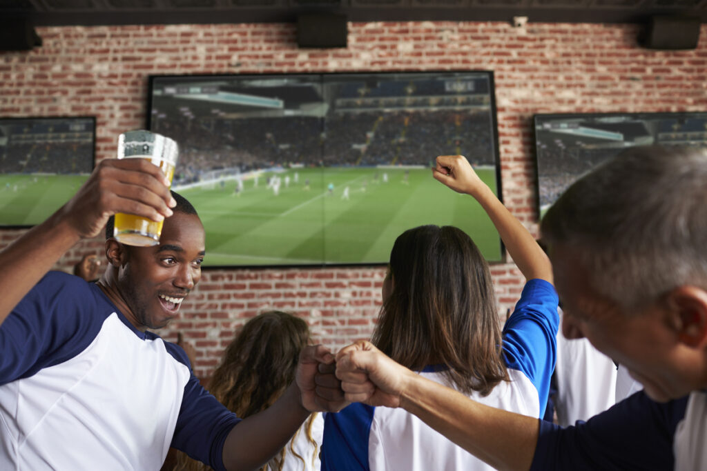Group of friends in a sports bar celebrating a soccer match as they fist-bump and cheer at big screens on a brick wall.