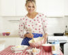 Smiling woman in a floral sweater and blue polka-dot apron mixes batter in a pink bowl at a bright kitchen counter with baking ingredients on a red-and-white checkered tablecloth.