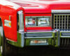 Front end of a red vintage car with a chrome bumper and rectangular headlights on a grassy background.