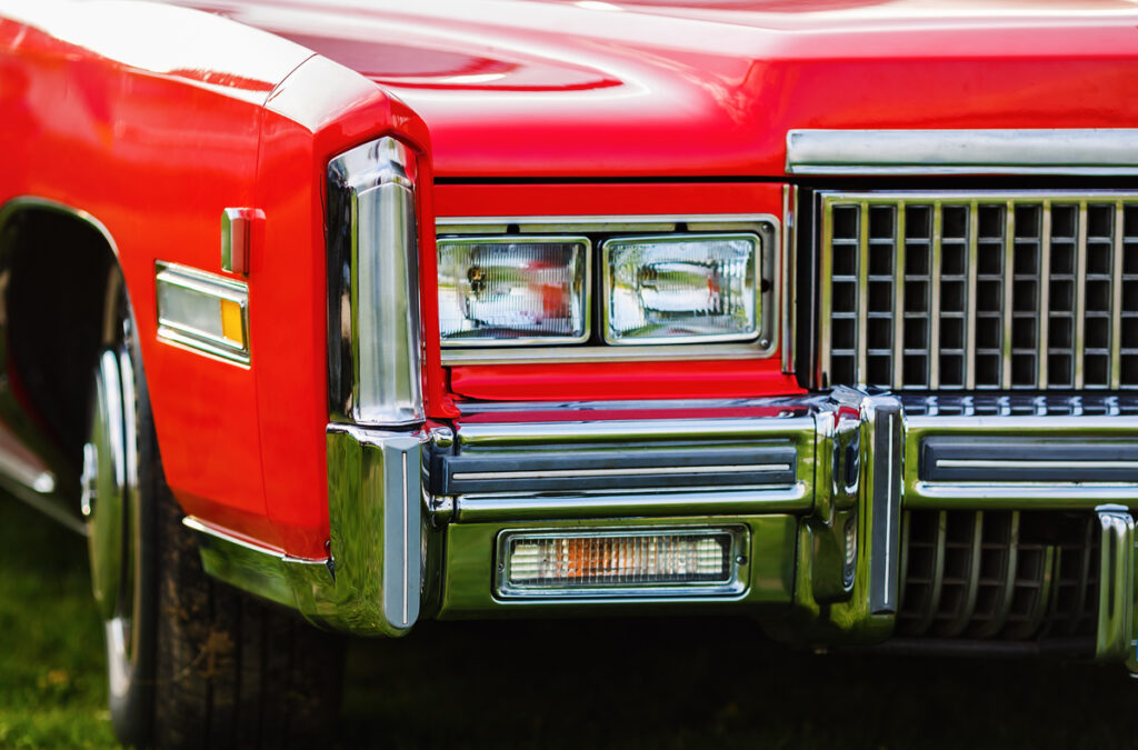 Front end of a red vintage car with a chrome bumper and rectangular headlights on a grassy background.