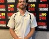 Man in a white polo shirt with folded hands stands center in front of a FM99 WNOR 'Rumble in the Morning' backdrop banner.