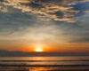Sunset over the ocean with orange horizon, golden clouds, and reflections on wet sandy shore.