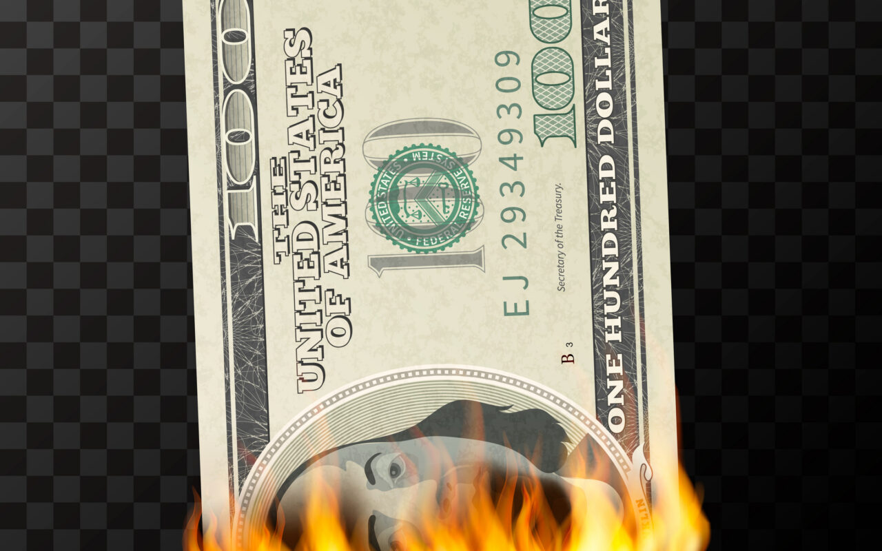 A close-up of a U.S. $100 bill burning at the bottom, flames licking the edge against a dark checkered background.