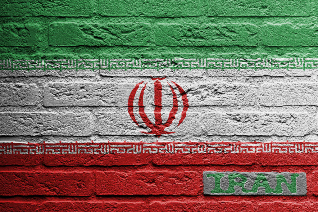 Iran flag painted on a rough brick wall, with green white red stripes and central emblem; 'IRAN' label on a gray badge in the corner.