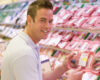 Smiling man in a light polo shirt holds a packaged raw meat in a supermarket meat aisle.