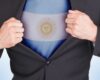 Man in a black suit pulls open his shirt to reveal an Argentina flag on his chest, signaling national pride or patriotism in a business context.