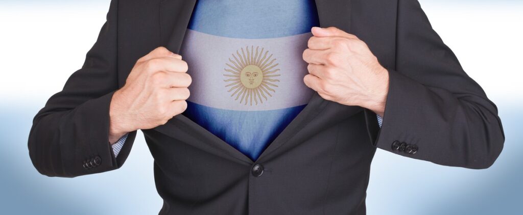 Man in a black suit pulls open his shirt to reveal an Argentina flag on his chest, signaling national pride or patriotism in a business context.