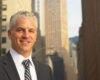 Portrait of a smiling businessman in a pinstripe suit and striped tie, outdoors on a city street with tall buildings in the background.