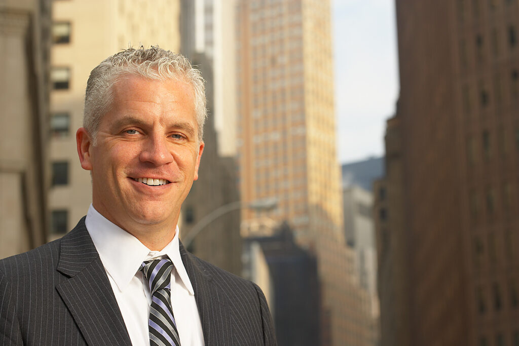Portrait of a smiling businessman in a pinstripe suit and striped tie, outdoors on a city street with tall buildings in the background.