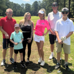 Group photo of six people posing on a golf course with clubs, smiling in sunny weather, casually dressed for golf.