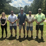 Five adults stand in a line on a sunny golf course, each holding a golf club and posing for a group photo, trees in the background.