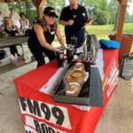 Woman in a black outfit and cap arranges trophies and a championship belt on a red-clothed table at an outdoor FM99 Rocks event; a man with a microphone stands nearby under a wooden pavilion.