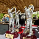 Set of silver golfer figurine trophies on a red base, with plaques reading '1st PLACE 2026 Rumble in the Morning Who's Your Caddy Summer Edition,' under a wooden pavilion.