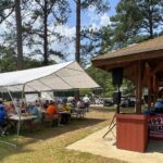People seated at long picnic tables under a white canopy at an outdoor event, eating and socializing.